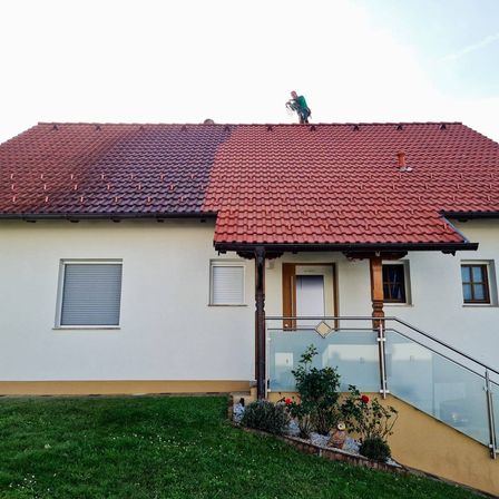 Ein Mann auf einem roten Dach reinigt die Ziegel, während ein Haus mit einer Glasbrüstung, weißen Wänden und Fenstern darunter sichtbar ist.