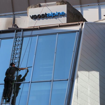 Ein Arbeiter auf einer Leiter reinigt die Glasfenster eines Gebäudes mit dem Schild Voestalpine darüber.