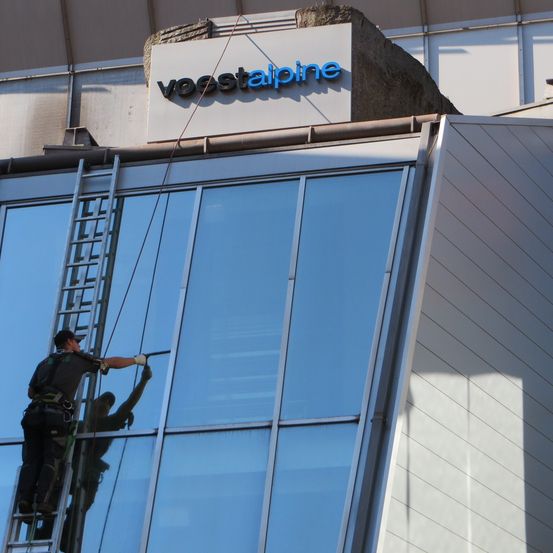 Ein Arbeiter auf einer Leiter reinigt die Glasfenster eines Gebäudes mit dem Schild Voestalpine darüber.