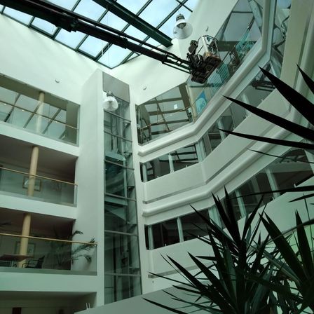Ein Innenansicht eines modernen Gebäudes mit Glaswänden und einer Glasdecke. Eine Person nutzt einen Hebebühne, um die Fenster zu reinigen.