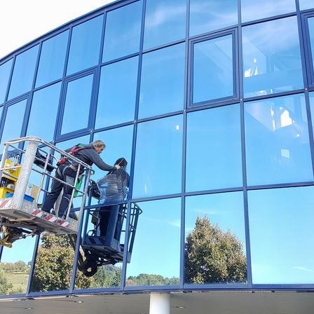 Zwei Personen reinigen Fenster eines Gebäudes. Eine Person ist auf einem Lift, die andere im Lift. Sie tragen Schutzausrüstung. Das Gebäude hat blaue Fensterrahmen.
