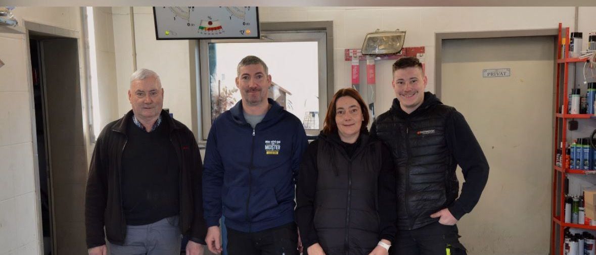 Vier Personen, darunter zwei Männer und zwei Frauen, stehen in einer Garage mit einem Hund. Eine Frau hält die Leine eines Hundes. Dahinter befindet sich ein Regal mit Flaschen und anderen Gegenständen.