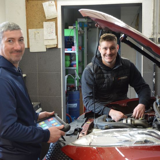 Zwei Männer in einer Garage, einer hält ein Tablet, der andere arbeitet am Automotor. Dahinter befindet sich ein Bulletin Board, ein Regal und ein blauer Fass.