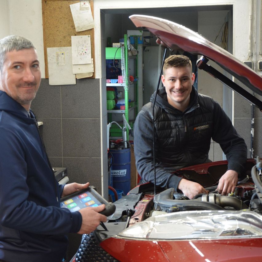 Zwei Männer in einer Garage, einer hält ein Tablet, der andere arbeitet am Automotor. Dahinter befindet sich ein Bulletin Board, ein Regal und ein blauer Fass.