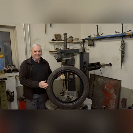 Ein Mann steht in einer Garage, lächelt und steht neben einem Reifen, der auf einer Maschine montiert ist. Werkzeuge und Ausrüstung sind im Hintergrund sichtbar.