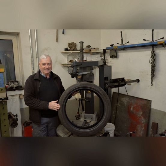 Ein Mann steht in einer Garage, lächelt und steht neben einem Reifen, der auf einer Maschine montiert ist. Werkzeuge und Ausrüstung sind im Hintergrund sichtbar.