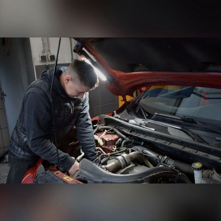 Ein Mann in einer schwarzen Jacke arbeitet in einer Garage an einem Automotor. Die Motorhaube des Autos ist geöffnet, und der Mann wird von einem Licht beleuchtet.