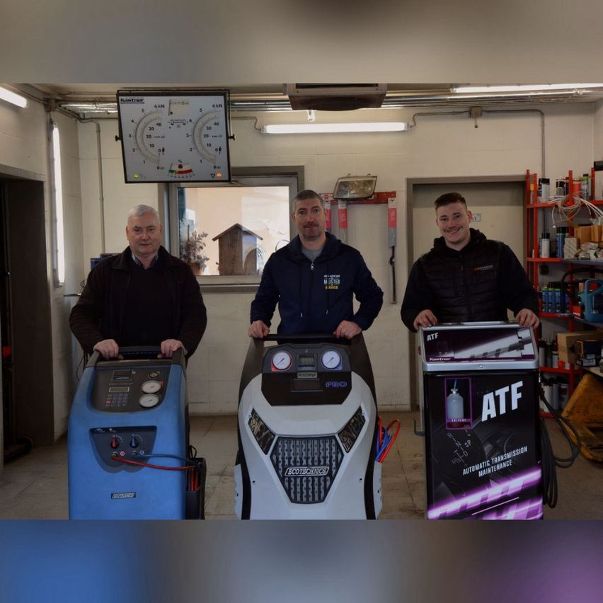 Drei Männer in einer Garage posieren neben Ausrüstung. Ein Mann hält eine blaue Maschine, ein anderer eine schwarz-weiße Maschine und der dritte eine lila Maschine mit ATF darauf.