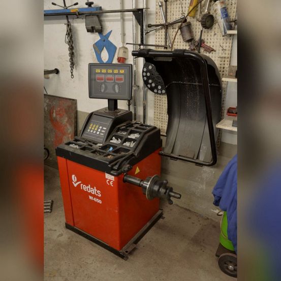Eine rote und schwarze Radausrichtungsmaschine, beschriftet mit 'redats W-650', steht in einer Werkstatt. Verschiedene Werkzeuge und Geräte hängen an der Wand dahinter.