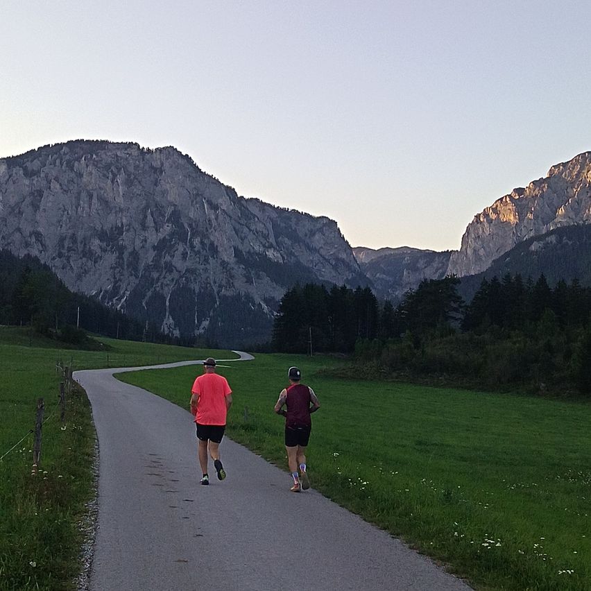 Two people are jogging on a winding path in a grassy field with mountains in the background.