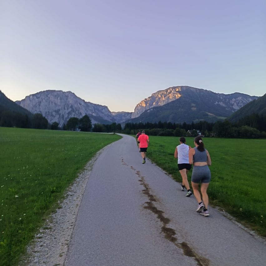 Three people are jogging on a gravel path in a grassy field with mountains in the background.