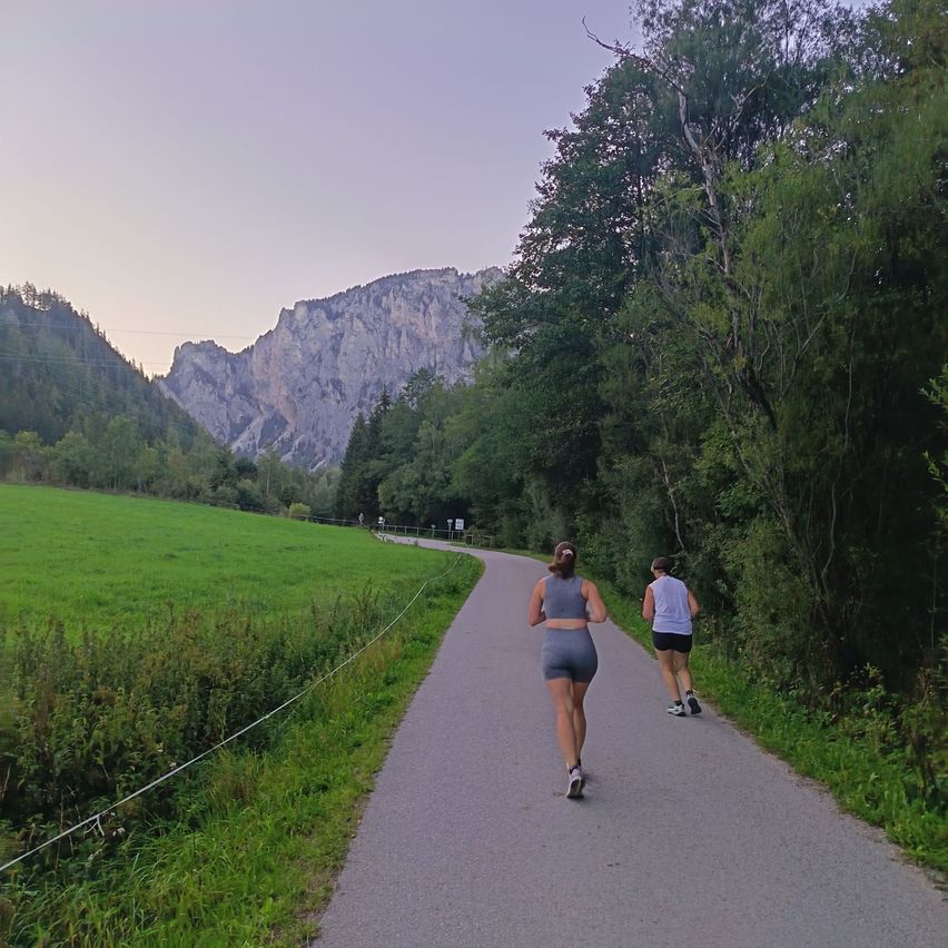 Two women jog on a winding path through a lush green valley, with mountains in the distance.
