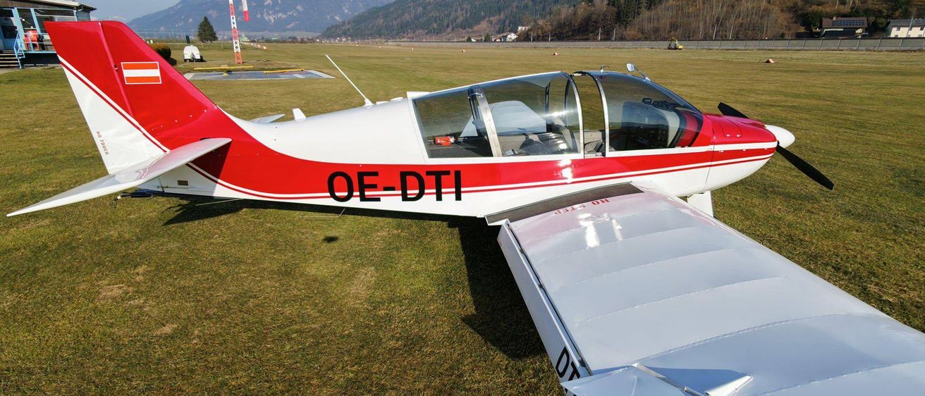 Ein kleines rotes und weißes Flugzeug mit der Kennung OE-DTI steht auf dem Gras eines Flugplatzes mit verschneigten Bergen im Hintergrund.