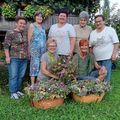 Sieben Frauen stehen in einem Garten, lächeln und posieren für ein Foto mit Blumenkörben. Die Frau in der Mitte hält einen großen Blumenstrauß. Hinter ihnen schmücken Pflanzen und Blumen den Garten.
