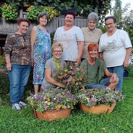 Sieben Frauen stehen in einem Garten, lächeln und posieren für ein Foto mit Blumenkörben. Die Frau in der Mitte hält einen großen Blumenstrauß. Hinter ihnen schmücken Pflanzen und Blumen den Garten.