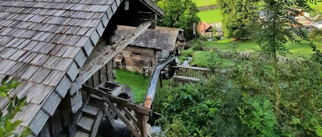 Eine Vogelperspektive auf ein Dorf mit einem rustikalen Holzhaus und Wasserrad in der Nähe einer grünen Wiese und eines Waldes unter einem bewölkten Himmel.