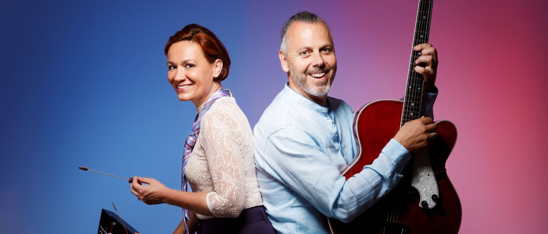 Ein Mann und eine Frau sitzen auf Hockern mit Musikinstrumenten. Die Frau hält einen Dirigentenstab und eine Zither. Der Mann hält eine Gitarre. Sie scheinen beide zu lächeln.