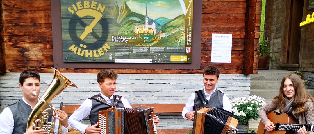 Four musicians sit on benches playing instruments outside a wooden building with a sign. One plays the tuba, two play accordions, and one plays the guitar.