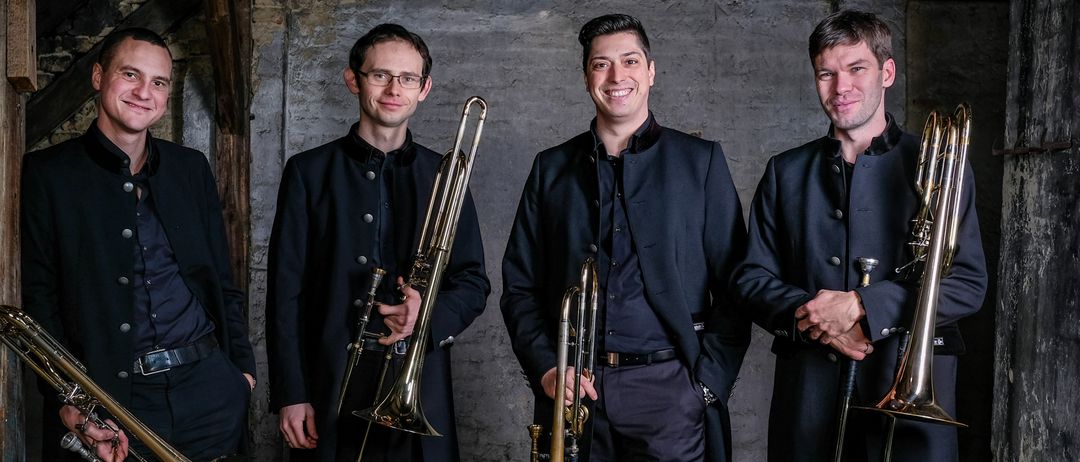 Four men in formal attire stand in a room, smiling and posing for a photo while holding trombones. The room has wooden beams and brick walls.