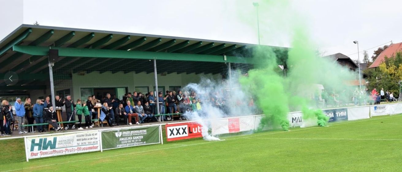 Ein Fußballfeld mit grünem Rauch, der aus dem Boden aufsteigt, möglicherweise von einem Feuerwerkskörper. Zuschauer beobachten von den Tribünen aus. Banner mit Werbung und Text sind an der Seite sichtbar.
