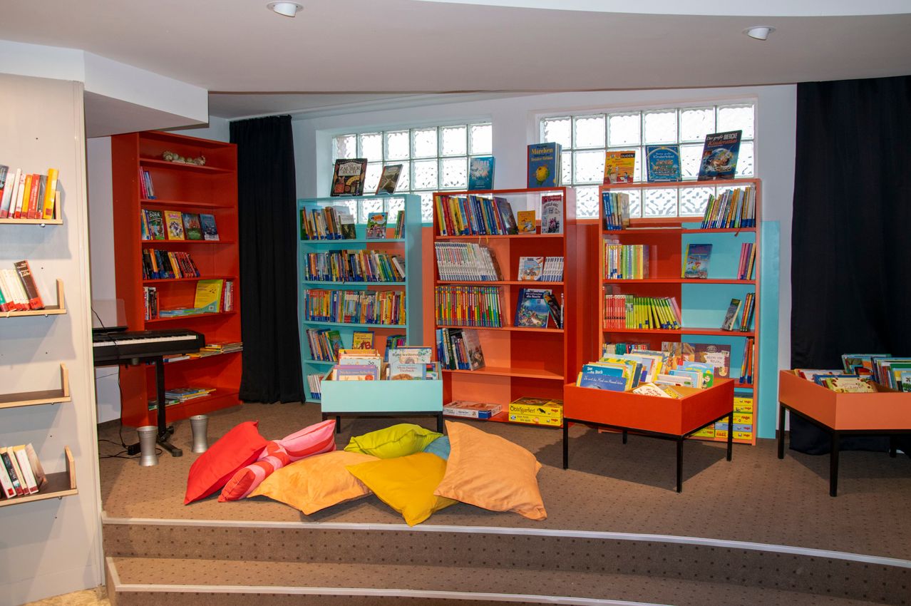 Ein farbenfroher Kinder-Lesebereich mit leuchtend orangefarbenen und türkisfarbenen Bücherregalen, gefüllt mit Büchern, weichen Kissen auf dem Boden, einem kleinen Klavier und einem Vorhang im Hintergrund.