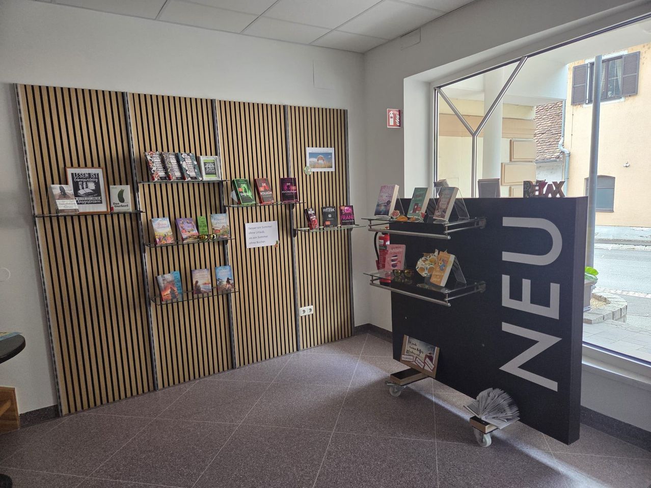 Ein leerer Raum mit Holzregalen, die Bücher präsentieren, und einem großen Schild, das 'NEU' auf der rechten Seite liest. Der Raum hat einen gefliesten Boden und ein Glasfenster auf der rechten Seite.