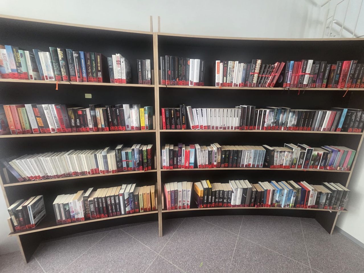 Two wooden bookshelves filled with numerous books, mostly fiction, arranged in rows with red ribbons marking the top of each book.
