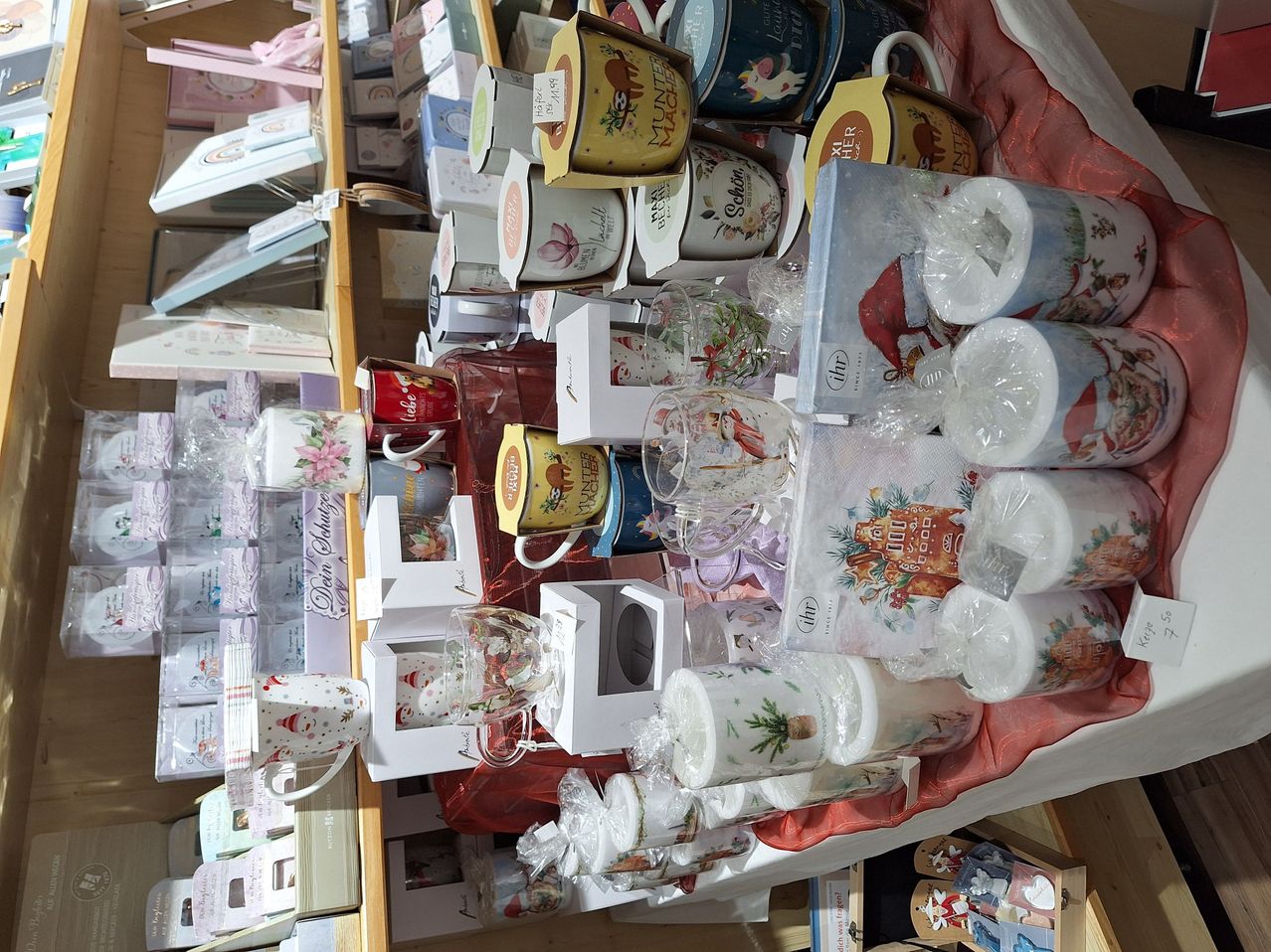 Eine Ausstellung von weihnachtlich gestalteten Tassen und Gläsern, einige in Plastik verpackt, auf einem Holzregal und einem Tisch angeordnet, mit verschiedenen anderen Produkten im Hintergrund.