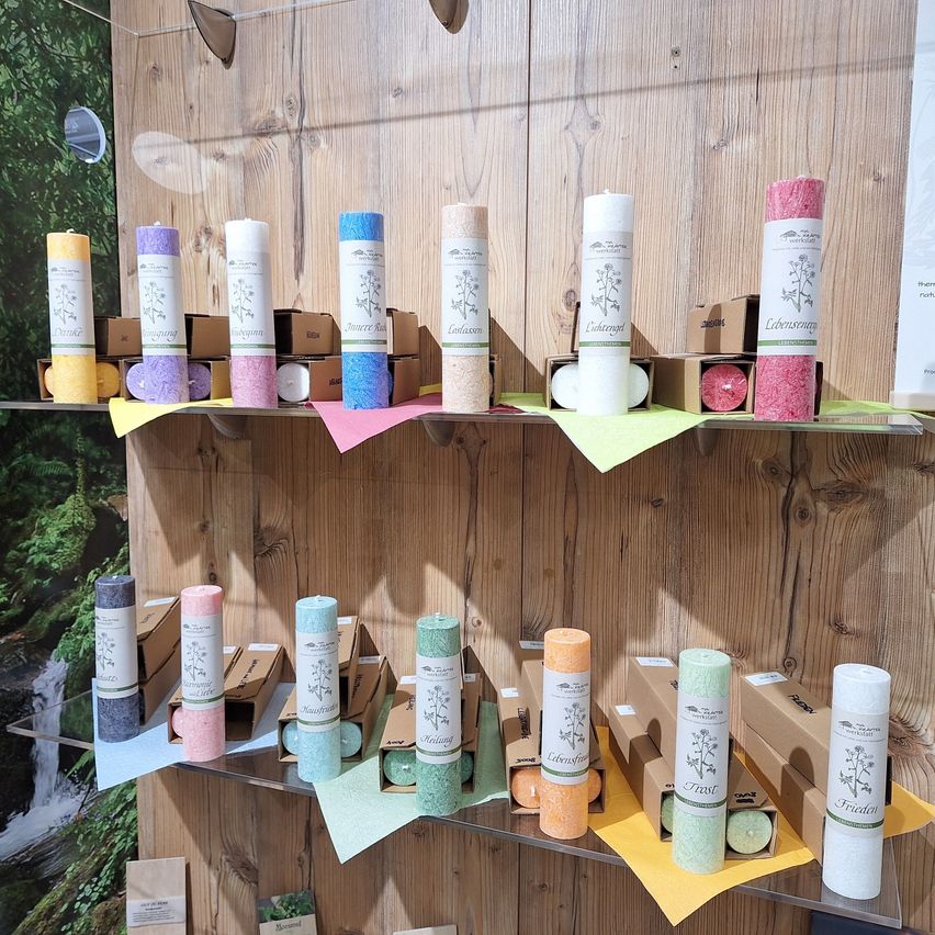 Ein Holzregal zeigt verschiedene farbige Kerzen, jede in einem Karton, in zwei Reihen auf einem Regal an einer Holzwand angeordnet.