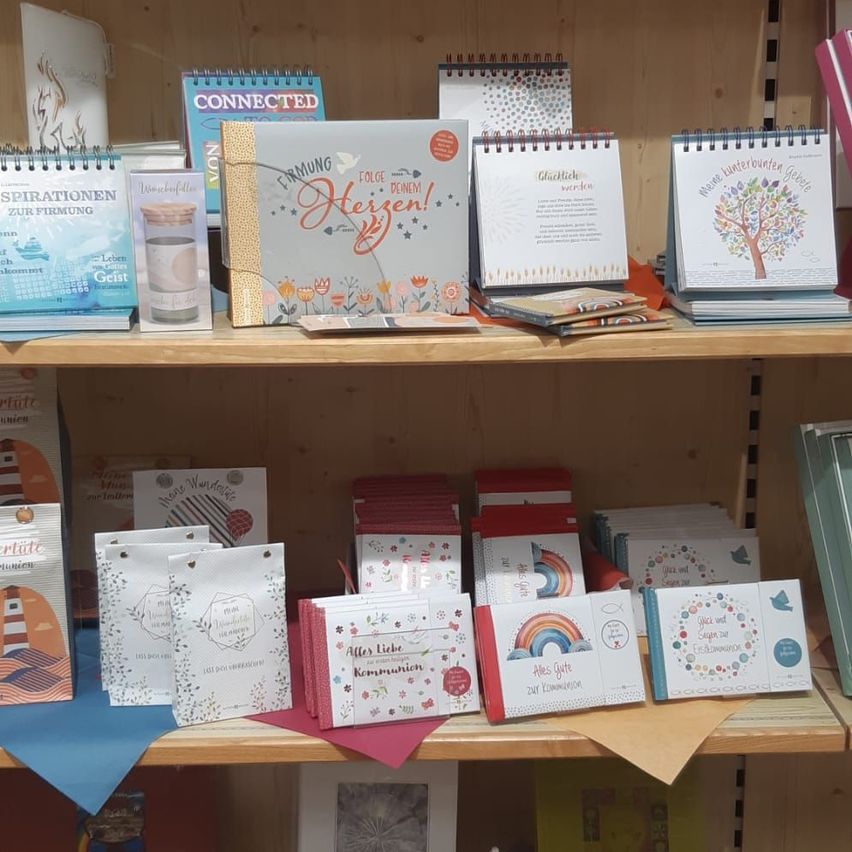 Ein hölzerne Regalplatte zeigt verschiedene Bücher und Broschüren. In der obersten Reihe befinden sich Kalender und ein Buch mit dem Titel 'Ermutigte'. Darunter sind Karten und kleine Bücher. Die Bücher haben farbenfrohe Umschläge mit Illustrationen und Text.