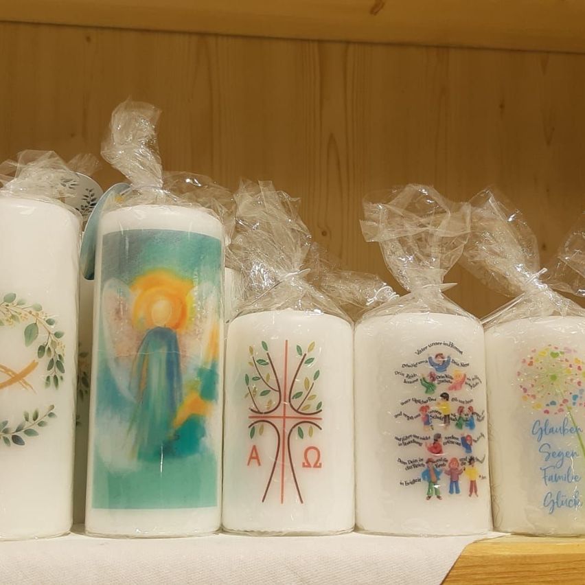Ein Holzregal hält sechs weiße Kerzen in Plastik. Jede Kerze zeigt verschiedene Designs: einen Engel, ein Kreuz, einen Baum und mehr. Sie sind ordentlich angeordnet und in Plastik eingewickelt.