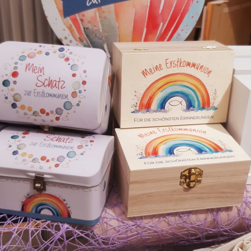 Drei Kisten auf einem Tisch. Die oberste Kiste hat die Aufschrift 'Mein Schatz zur Erstkommunion'. Die mittlere Kiste zeigt einen Regenbogen und hat die Aufschrift 'Meine Erstkommunion'. Die unterste Kiste hat die Aufschrift 'Schatz zur Erstkommunion'.