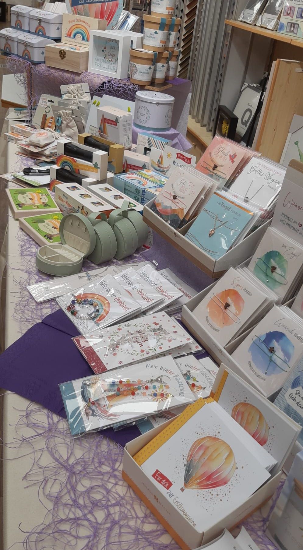 Ein Tisch zeigt eine Auswahl an Geschenkartikeln, einschließlich Karten, Schmuck und kleinen Schachteln, einige in Plastik. Bunte Karten mit Illustrationen und Text sind in verschiedenen Schachteln.