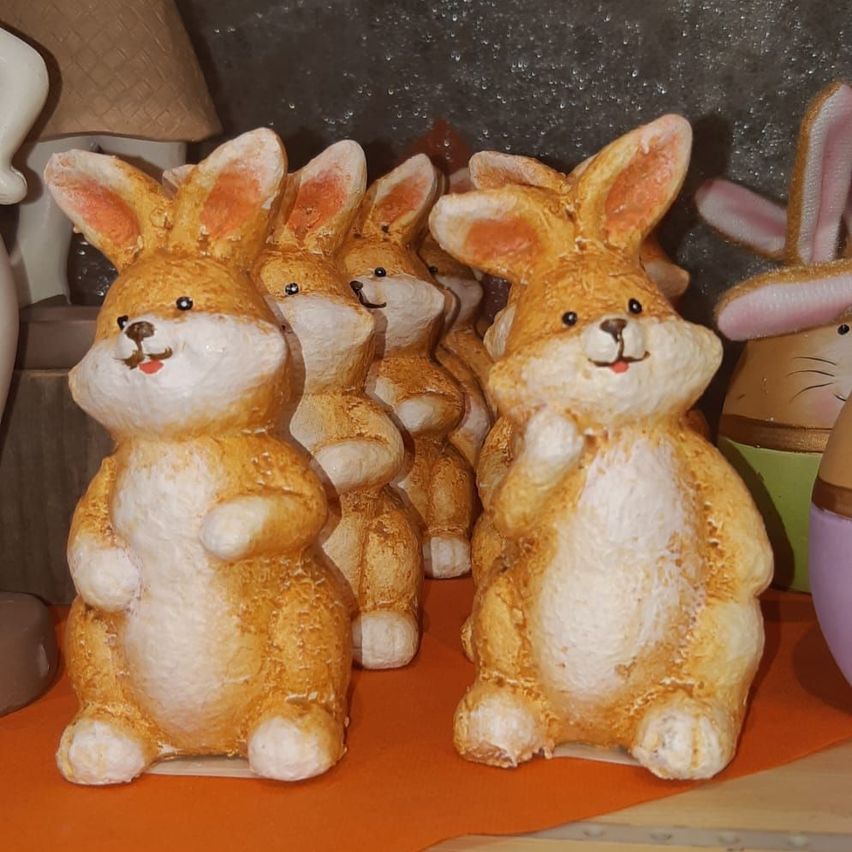 Eine Gruppe von Keramik-Hasenfiguren ist auf einer orangefarbenen Oberfläche angeordnet. Die Hasen haben braune und weiße Färbung mit rosa Ohren. Hinter ihnen befinden sich weitere Figuren, einschließlich eines weißen Hasen mit rosa Ohren.