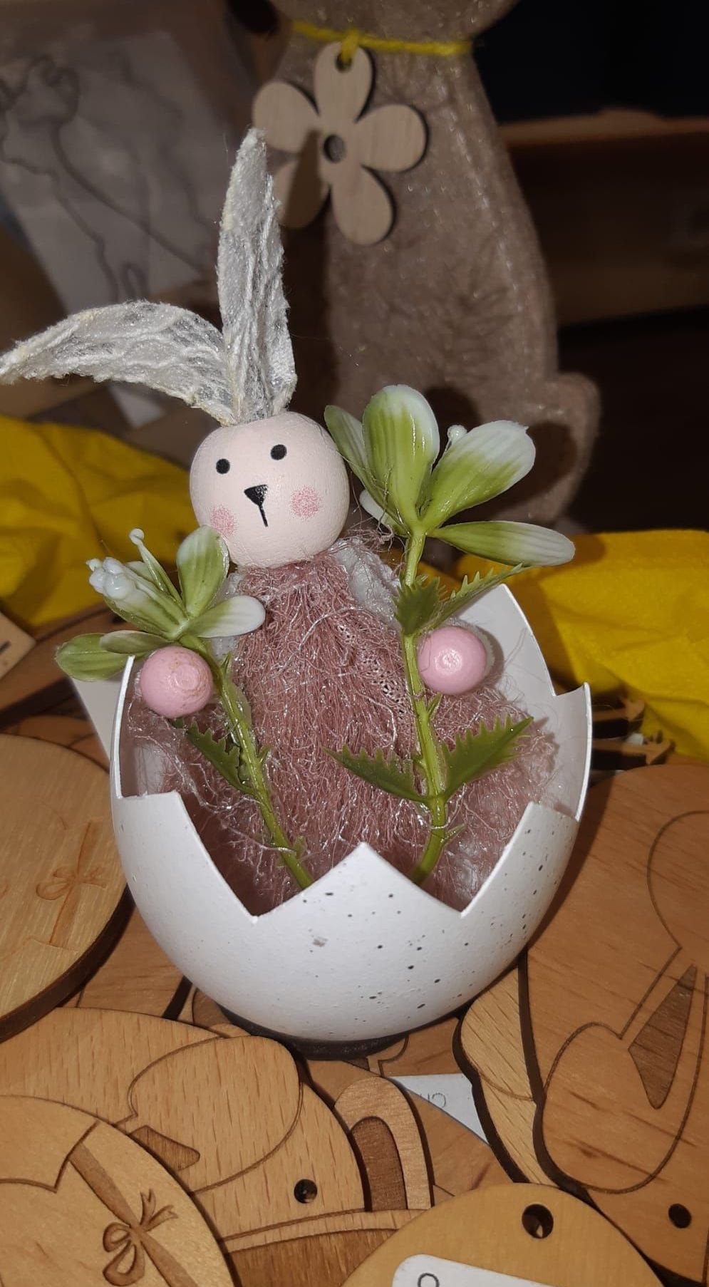 Eine Hasenfigur steckt in einem gecrackten Ei, umgeben von rosa und grünen Blumen. Der Hintergrund ist verschwommen mit einem gelben Tuch.