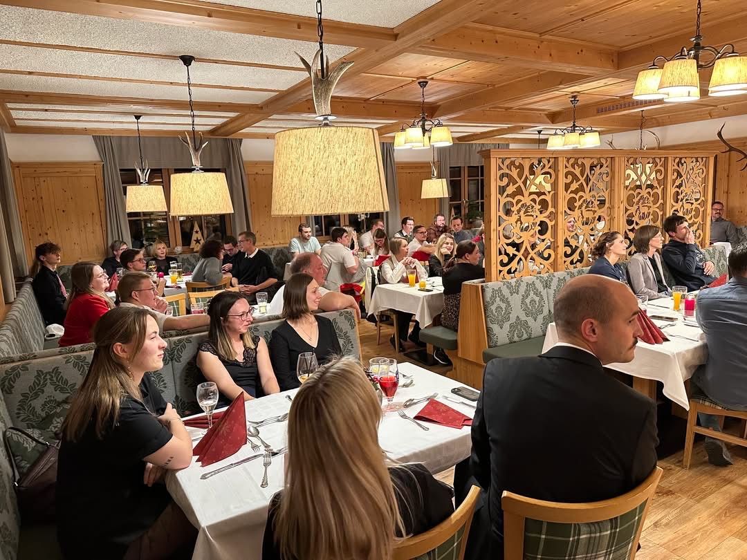 Ein Restaurant mit Holzdecken und Tischen mit roten Servietten. Mehrere Menschen sitzen und unterhalten sich. Einige Tische haben Weingläser und Getränke.