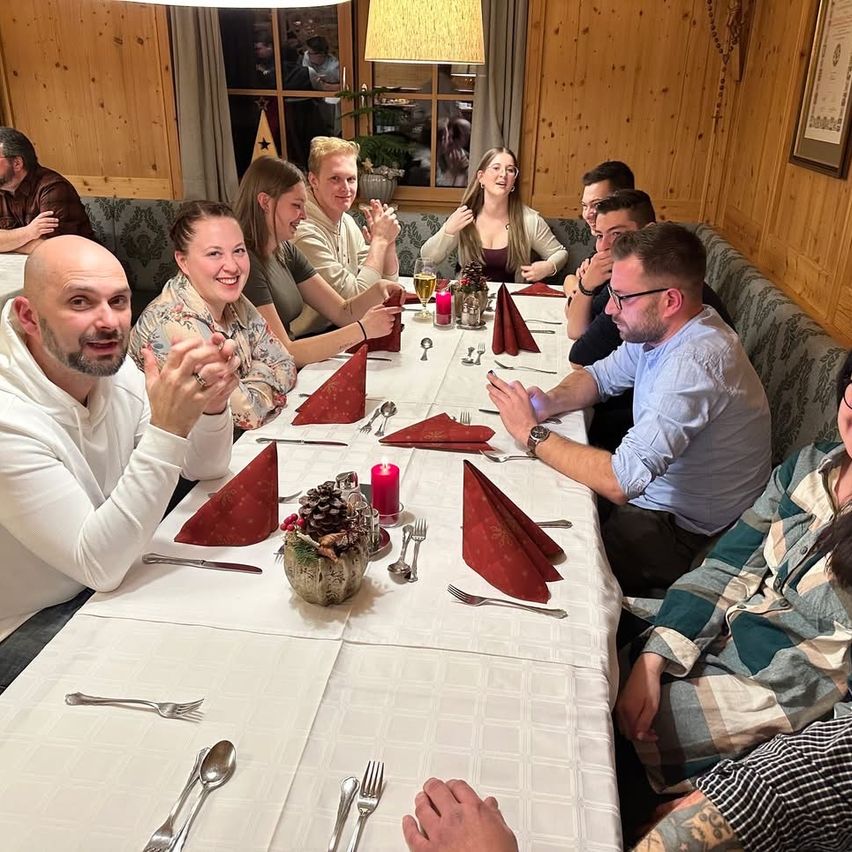 Eine Gruppe von Menschen sitzt in einem Restaurant an einem Tisch, lächelt und klatscht, mit Weinglasern, einer Kerze und Blumen auf dem Tisch.