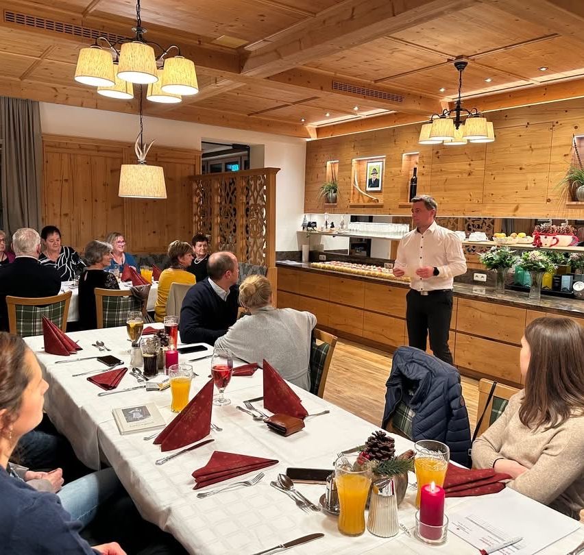 Eine Gruppe von Menschen sitzt um einen Tisch in einem Restaurant. Ein Mann steht vor ihnen an der Theke. Auf dem Tisch stehen Kerzen, Getränke und Essen. Das Restaurant hat Holzwände und -böden.