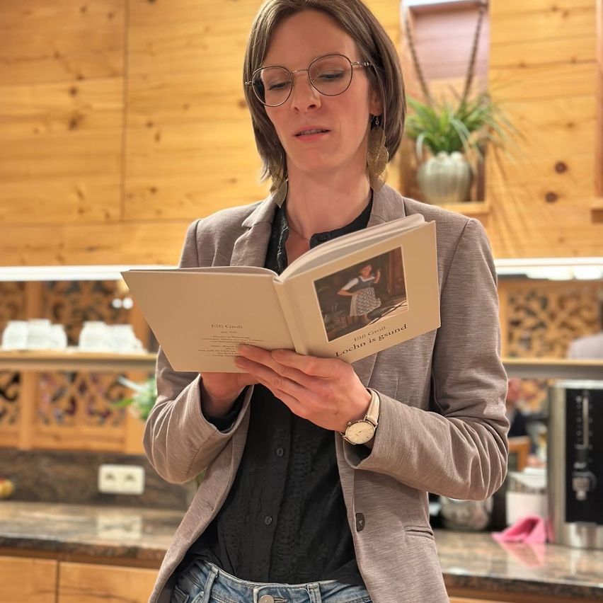 Eine Frau mit Brille und Blazer liest ein Buch mit dem Titel 'Loch is gesund' in einer Küche mit Holzschränken.