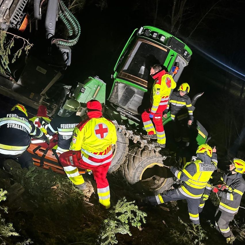 Eine Nachtrettung zeigt Feuerwehrleute in gelben und roten Uniformen, einer hält eine Trage, sie arbeiten daran, ein in Schlamm und Bäumen festsitzendes Fahrzeug zu befreien.