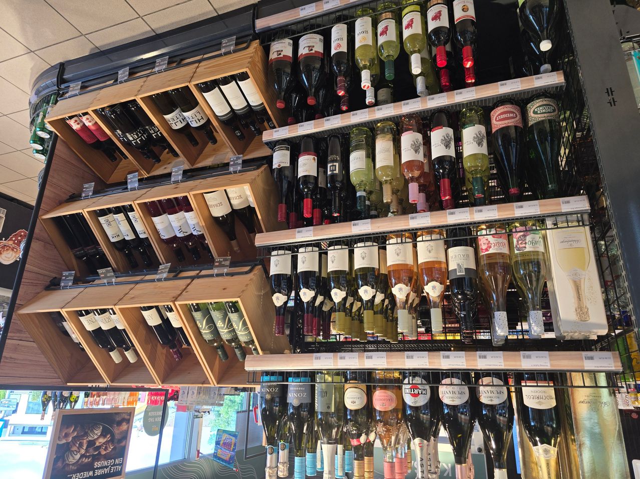 Ein Spirituosengeschäft zeigt eine große Auswahl an Weinflaschen auf Holzregalen und Metallständern, darunter Rot-, Weiß- und Roséweine, wobei Preisschilder an einigen Flaschen sichtbar sind.