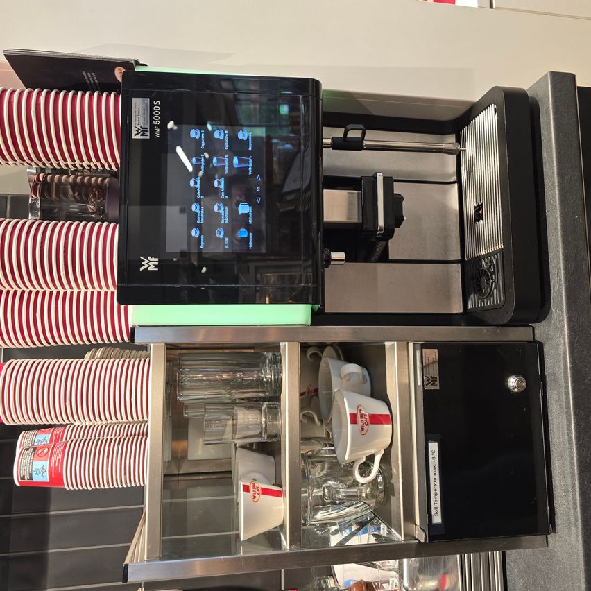 Eine schwarze Kaffeemaschine mit einem digitalen Display, das Getränkeoptionen anzeigt. Es hat einen Stapel roter und weißer Tassen auf der linken Seite und einen Glasschrank mit Tassen und Gläsern auf der rechten Seite.