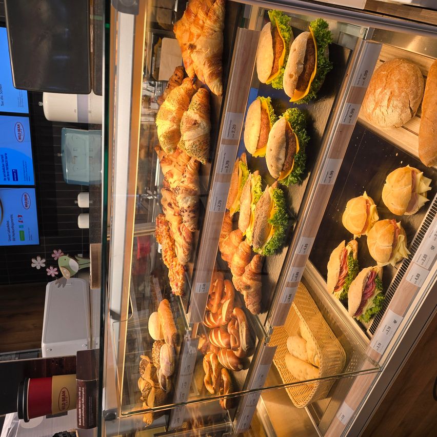 Eine Glasvitrine enthält verschiedene Gebäcksorten, darunter Croissants und Sandwiches. Die Croissants haben verschiedene Toppings, und die Sandwiches sind auf Tabletts mit Preisschildern platziert.