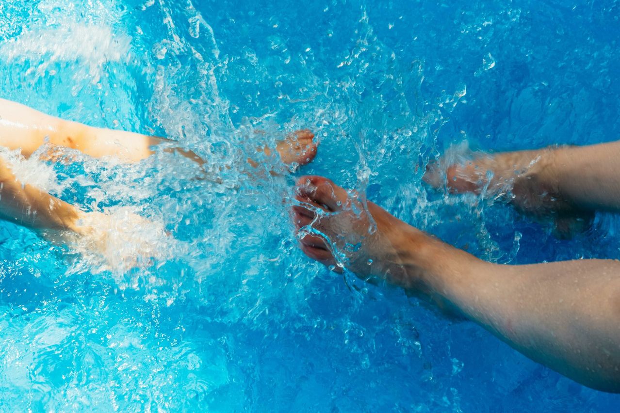 Ein Nahaufnahme von zwei nackten Füßen, die in einem Schwimmbad planschen, ein Fuß nach dem anderen greifend. Wassertropfen sind um die Füße herum sichtbar.