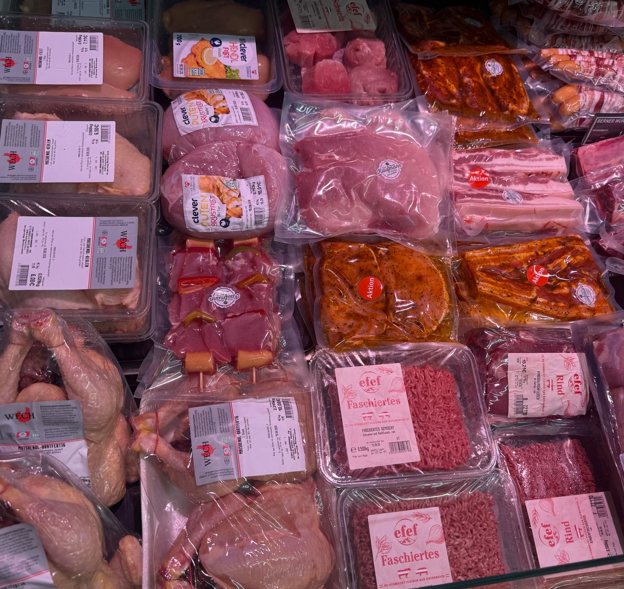 Verschiedene Fleisch- und Geflügelstücke sind in durchsichtigen Plastikbehältern ausgestellt, jeweils mit Etiketten, die Art und Gewicht angeben. Hühnchen, Rind und Schwein sind prominent vertreten.
