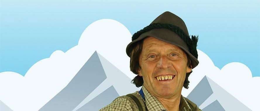 Ein lächelnder Mann in Hut und Rucksack steht vor einer Kulisse von Bergen und Hügeln. Er trägt eine blaue Schürze mit einem weißen Logo.