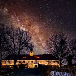 Ein Haus mit einem Glockenturm unter einem Sternenhimmel, die Milchstraße sichtbar. Das Gebäude ist hell beleuchtet und die Umgebung ist dunkel.