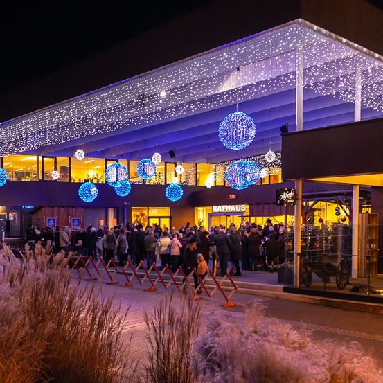 Eine Menschenmenge versammelt sich bei Nacht vor einem Gebäude, das mit blauen Lichtern und Ornamenten geschmückt ist. Das Gebäude hat ein modernes Design mit einer Glasfassade.