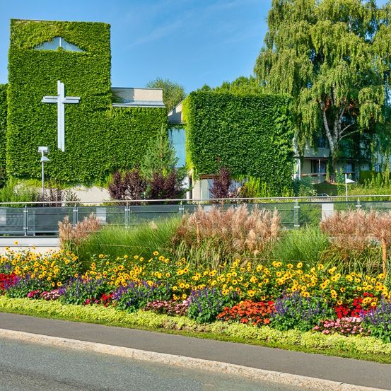 Eine Kirche mit Efeu-bewachsenen Mauern, einem Kreuz und einem Garten mit verschiedenen Blumen steht am Straßenrand. Der Himmel ist blau und klar.
