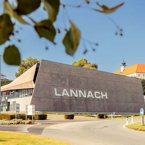 Ein großes Gebäude mit dem Wort Lannach darauf, ein blauer Himmel darüber, eine Straße davor und Bäume und Pflanzen an der Seite.
